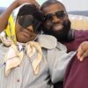Happy couple wearing sunglasses lounge on a couch, with the woman in a striped blouse and yellow scarf resting her head on the man’s shoulder.
