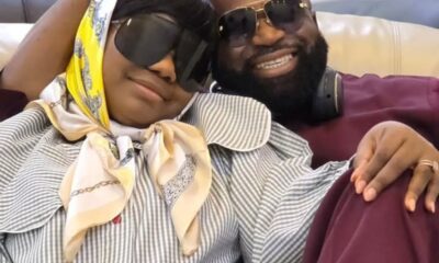 Happy couple wearing sunglasses lounge on a couch, with the woman in a striped blouse and yellow scarf resting her head on the man’s shoulder.