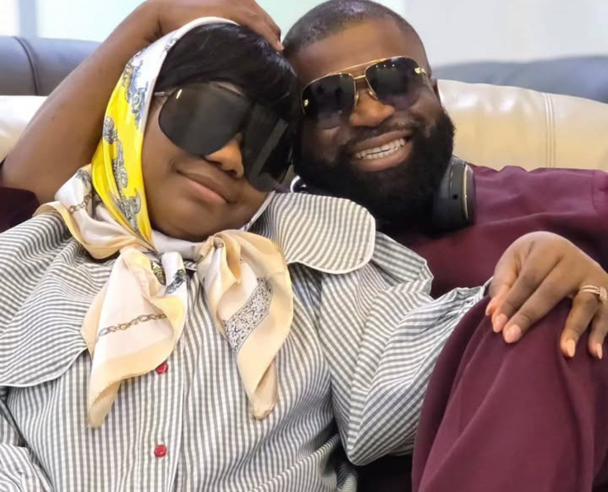 Happy couple wearing sunglasses lounge on a couch, with the woman in a striped blouse and yellow scarf resting her head on the man’s shoulder.