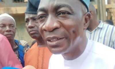 Close-up of a man in a white traditional outfit being interviewed, with a blue microphone featuring a world map logo in the foreground and onlookers behind him.