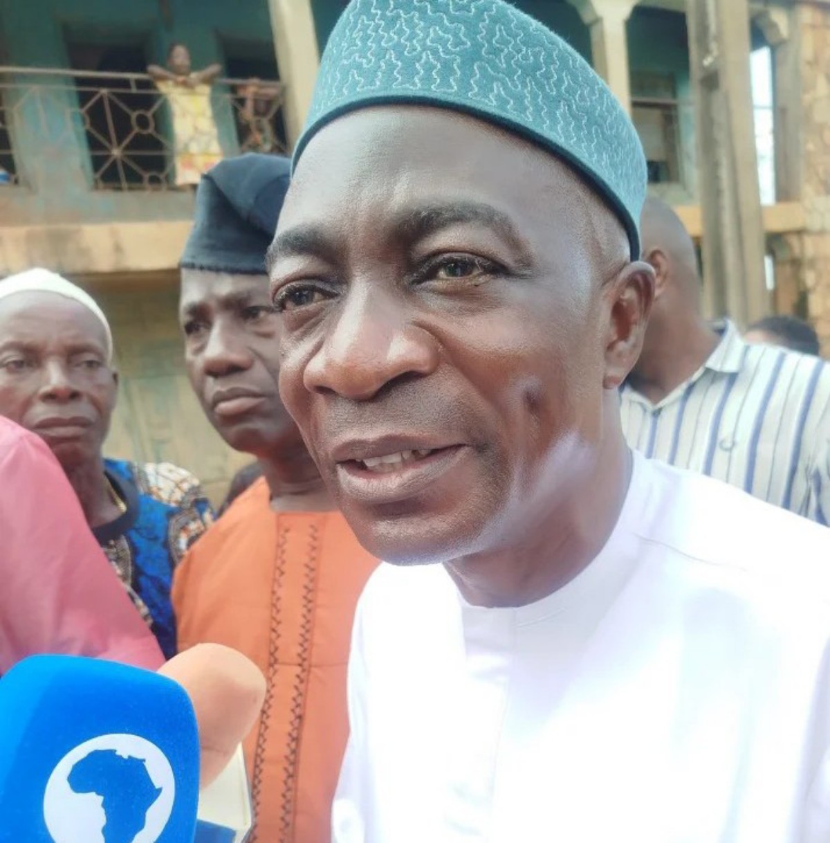 Close-up of a man in a white traditional outfit being interviewed, with a blue microphone featuring a world map logo in the foreground and onlookers behind him.