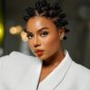Portrait of a woman with bantu knot hairstyle, wearing a white blazer and green clover jewelry, looking to the side in a studio setting.