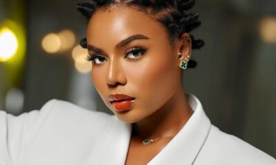 Portrait of a woman with bantu knot hairstyle, wearing a white blazer and green clover jewelry, looking to the side in a studio setting.