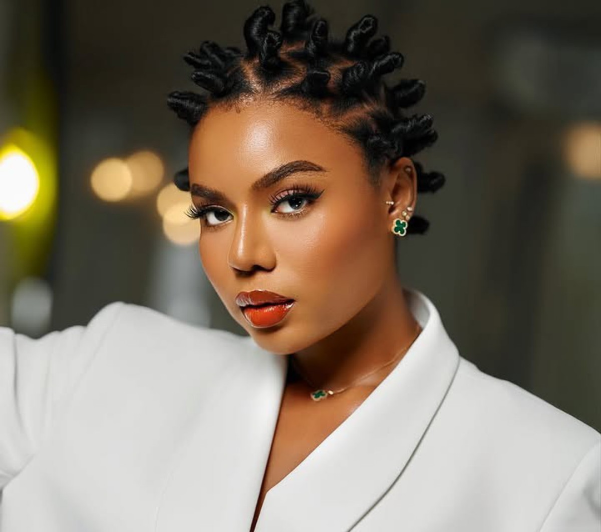 Portrait of a woman with bantu knot hairstyle, wearing a white blazer and green clover jewelry, looking to the side in a studio setting.