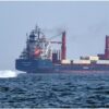 Large cargo ship on the open sea with a small speedboat ahead, suggesting a rescue or escort operation in progress.