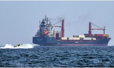 Large cargo ship on the open sea with a small speedboat ahead, suggesting a rescue or escort operation in progress.