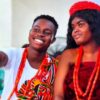 Two young people in colorful traditional attire, smiling and wearing red beaded necklaces at a cultural celebration.