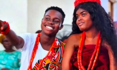 Two young people in colorful traditional attire, smiling and wearing red beaded necklaces at a cultural celebration.