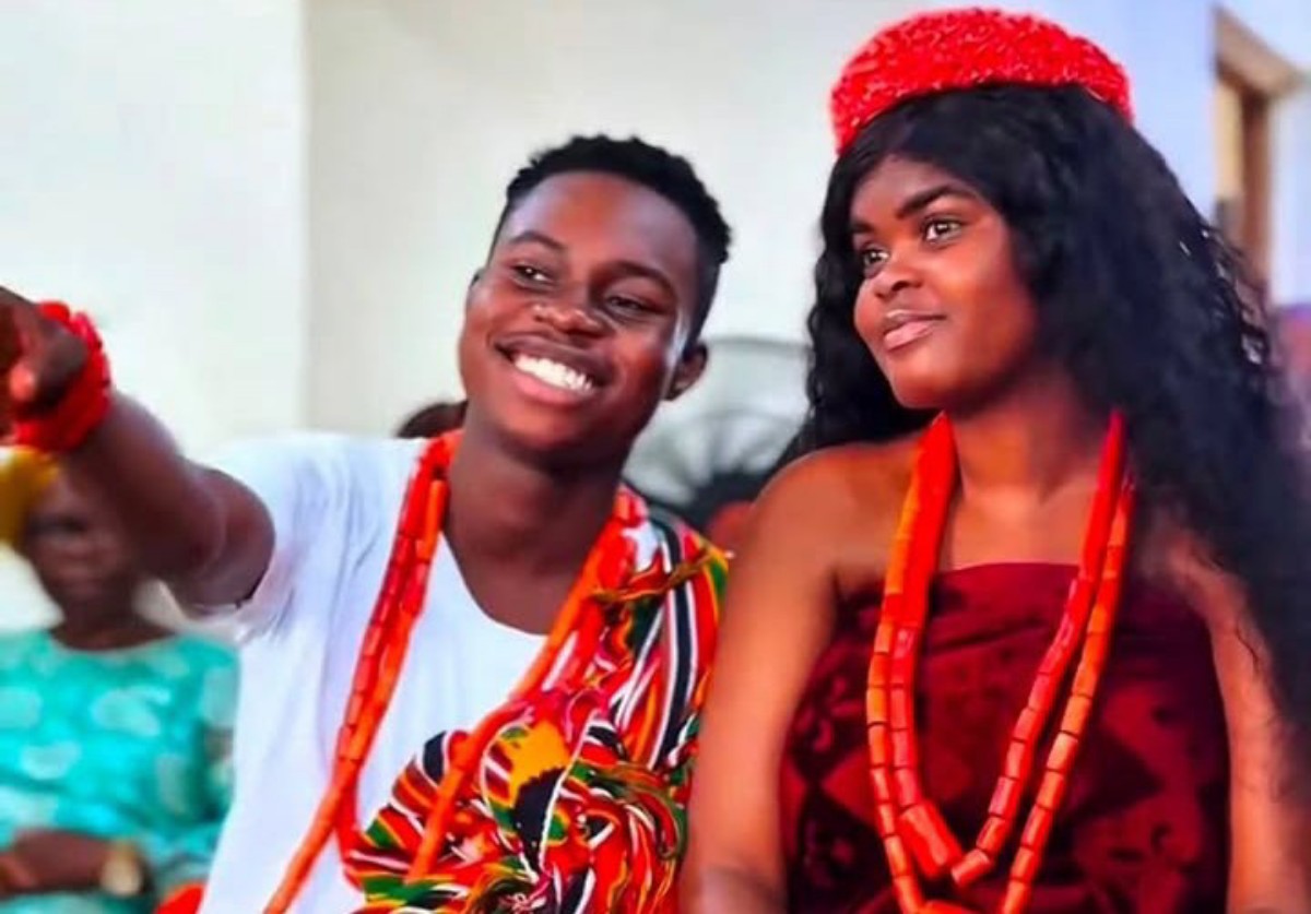 Two young people in colorful traditional attire, smiling and wearing red beaded necklaces at a cultural celebration.