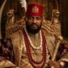 Portrait of a man in an ornate gold-embroidered robe wearing a red cap with a crest, seated on a gilded throne with a scepter and beaded jewelry.