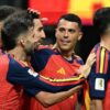Spanish soccer players in red-blue jerseys celebrate a goal, arms around each other in a group hug on the field