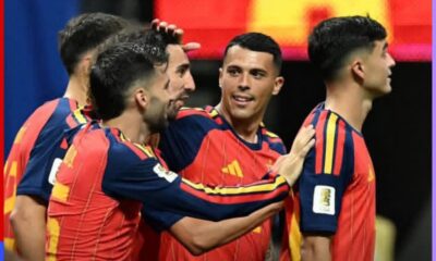 Spanish soccer players in red-blue jerseys celebrate a goal, arms around each other in a group hug on the field