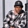 Man wearing a black cap that reads 'JESUS BOY' and a gray plaid jacket sits beside a black pole, looking at the camera.