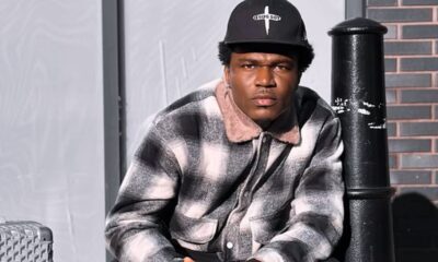 Man wearing a black cap that reads 'JESUS BOY' and a gray plaid jacket sits beside a black pole, looking at the camera.