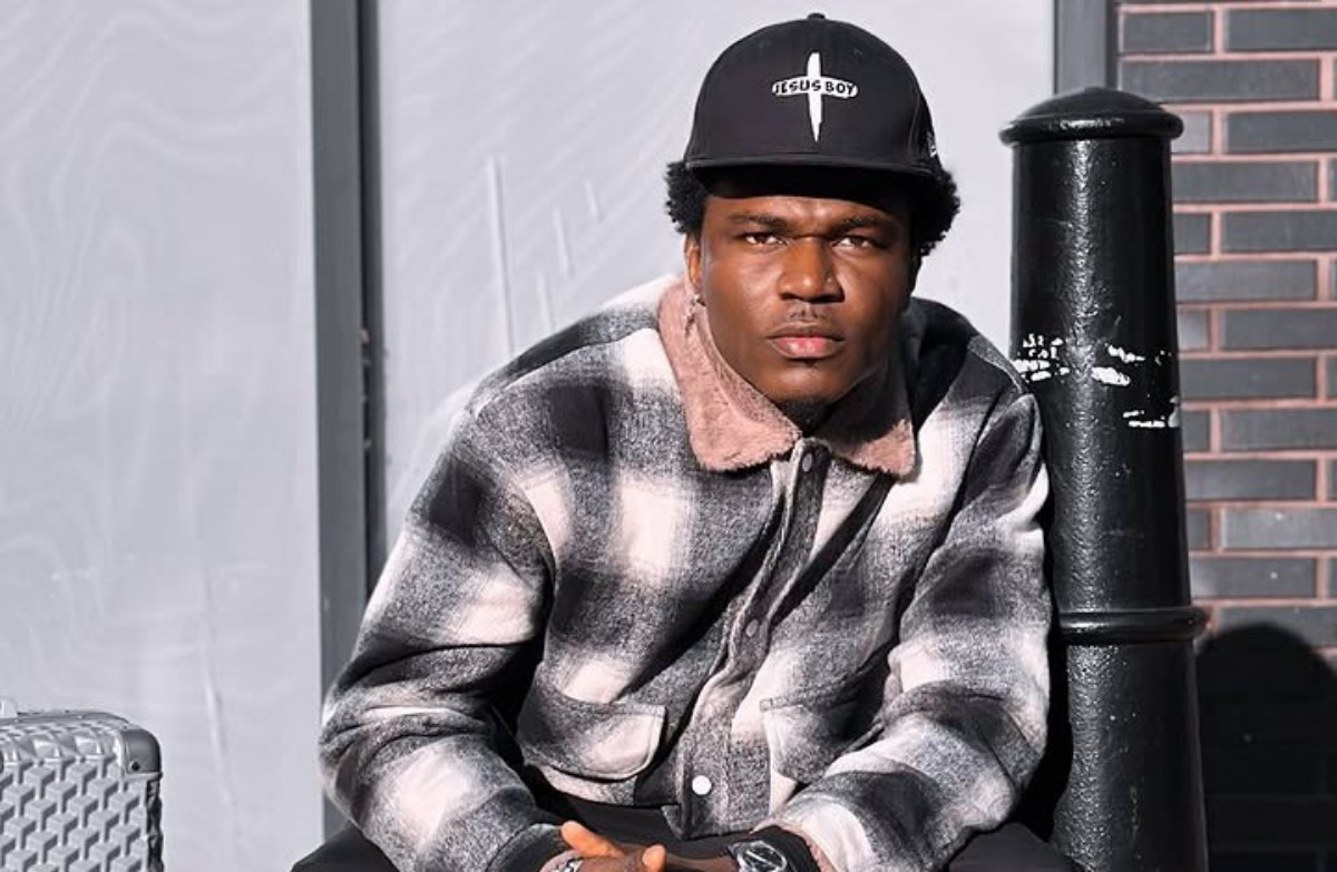Man wearing a black cap that reads 'JESUS BOY' and a gray plaid jacket sits beside a black pole, looking at the camera.