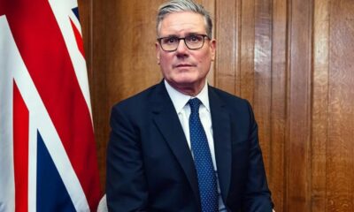 Man in a dark suit and blue tie sits indoors beside a partial UK flag; wood paneled wall in the background, wearing glasses.