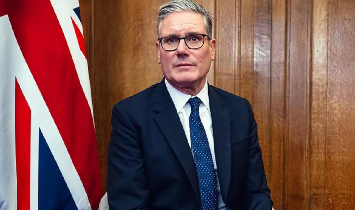 Man in a dark suit and blue tie sits indoors beside a partial UK flag; wood paneled wall in the background, wearing glasses.
