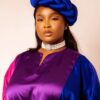 Woman wearing a color-block satin dress (purple, pink, blue) with a blue head wrap and a pearl choker against a neutral background.