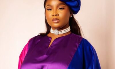 Woman wearing a color-block satin dress (purple, pink, blue) with a blue head wrap and a pearl choker against a neutral background.