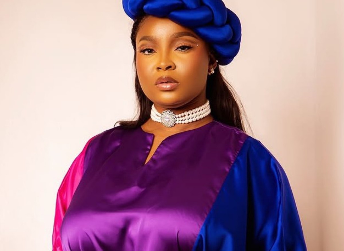 Woman wearing a color-block satin dress (purple, pink, blue) with a blue head wrap and a pearl choker against a neutral background.