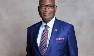 Portrait of a bald man in a navy suit, white shirt, red striped tie, glasses, and a red pocket square against a gray backdrop.