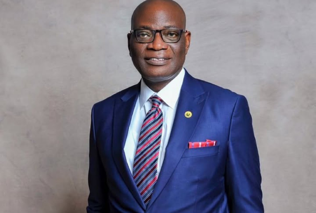 Portrait of a bald man in a navy suit, white shirt, red striped tie, glasses, and a red pocket square against a gray backdrop.