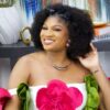 Smiling woman with a large natural afro wearing an off-shoulder white dress adorned with bright pink and green fabric flowers, posing beside a decorative table and books.