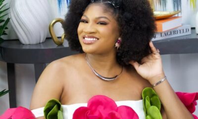 Smiling woman with a large natural afro wearing an off-shoulder white dress adorned with bright pink and green fabric flowers, posing beside a decorative table and books.