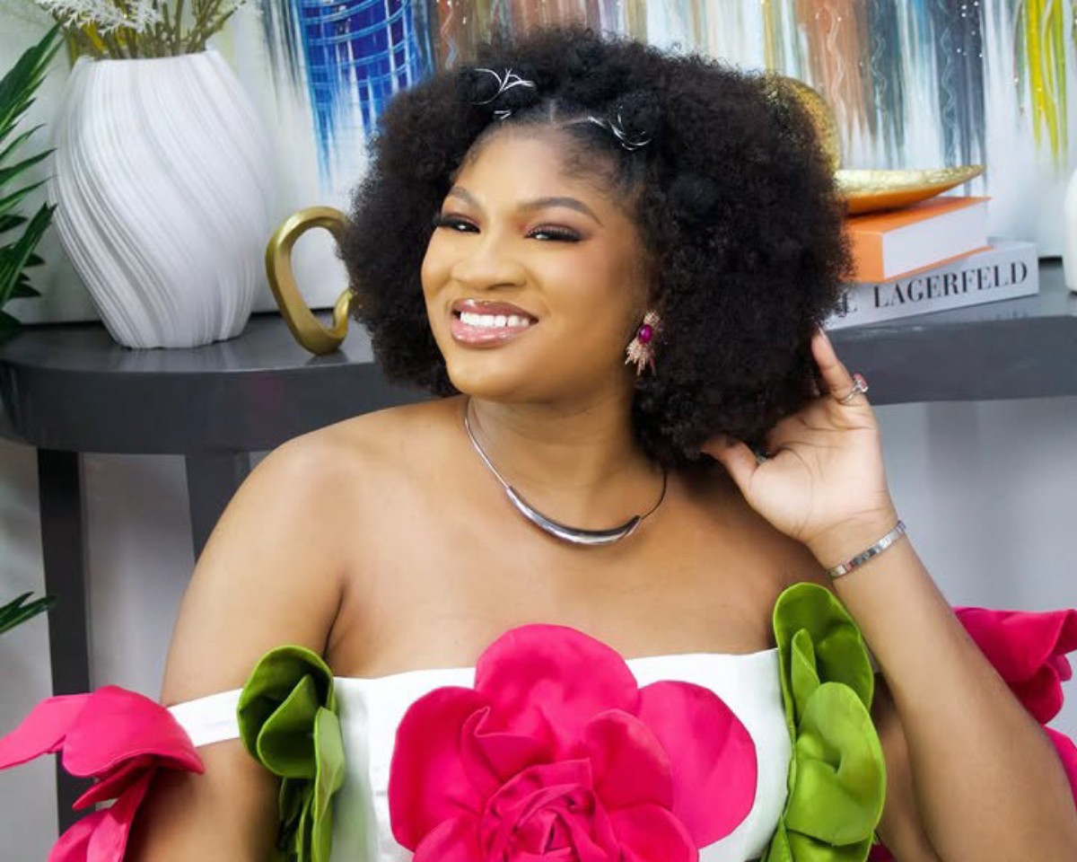 Smiling woman with a large natural afro wearing an off-shoulder white dress adorned with bright pink and green fabric flowers, posing beside a decorative table and books.