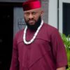 Man in maroon traditional attire (agbada) with a white beaded necklace and red fez, standing outdoors with greenery nearby.