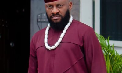 Man in maroon traditional attire (agbada) with a white beaded necklace and red fez, standing outdoors with greenery nearby.