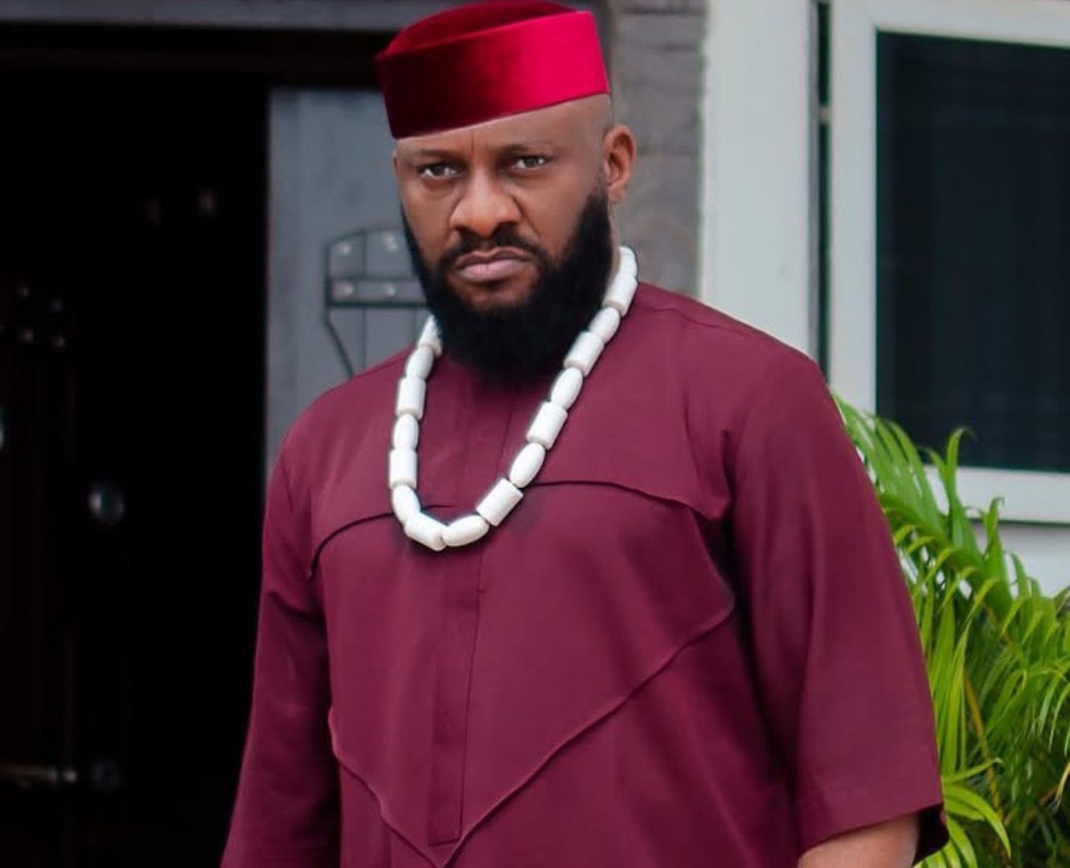 Man in maroon traditional attire (agbada) with a white beaded necklace and red fez, standing outdoors with greenery nearby.