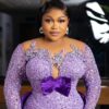 Woman in a lavender, beaded gown with a large purple bow, posing for a photo with a soft smile and elegant jewelry.