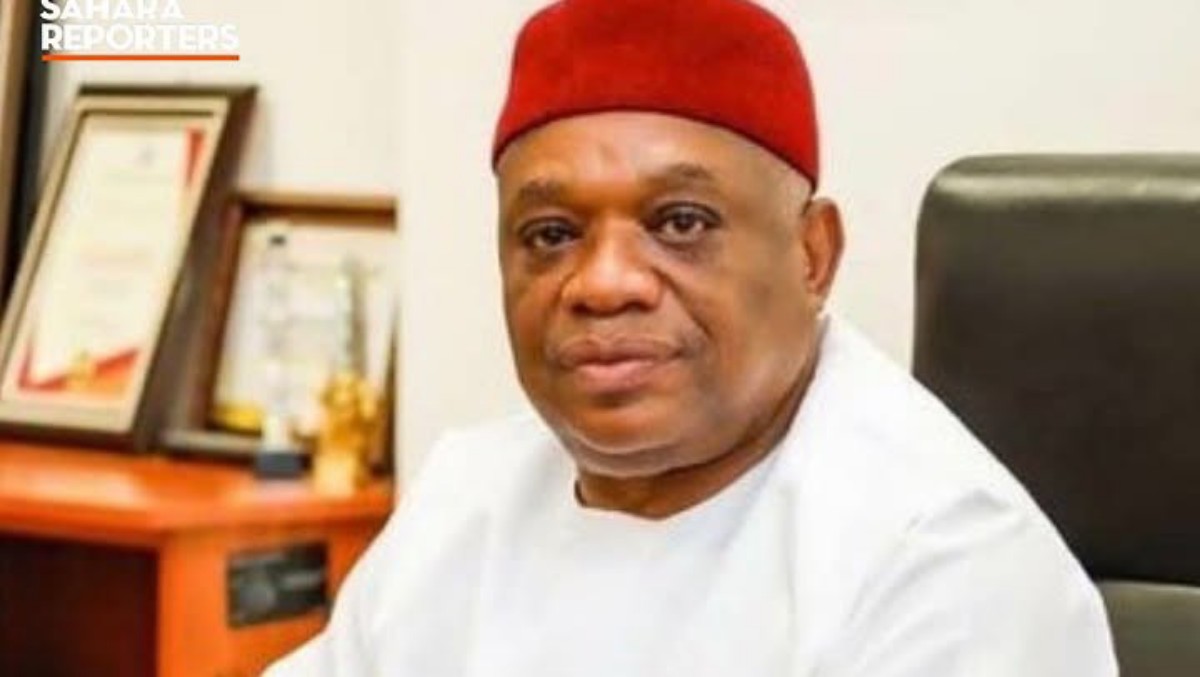 Portrait of a man in a white traditional outfit and red cap seated at a desk in an office, with awards in the background.