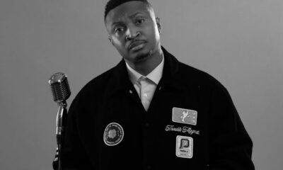 Black-and-white photo of a man in a patched jacket standing beside a vintage microphone, looking at the camera.