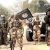 Group of armed militants in desert camouflage beside an armored vehicle, one raising a flag.