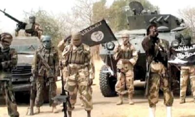 Group of armed militants in desert camouflage beside an armored vehicle, one raising a flag.