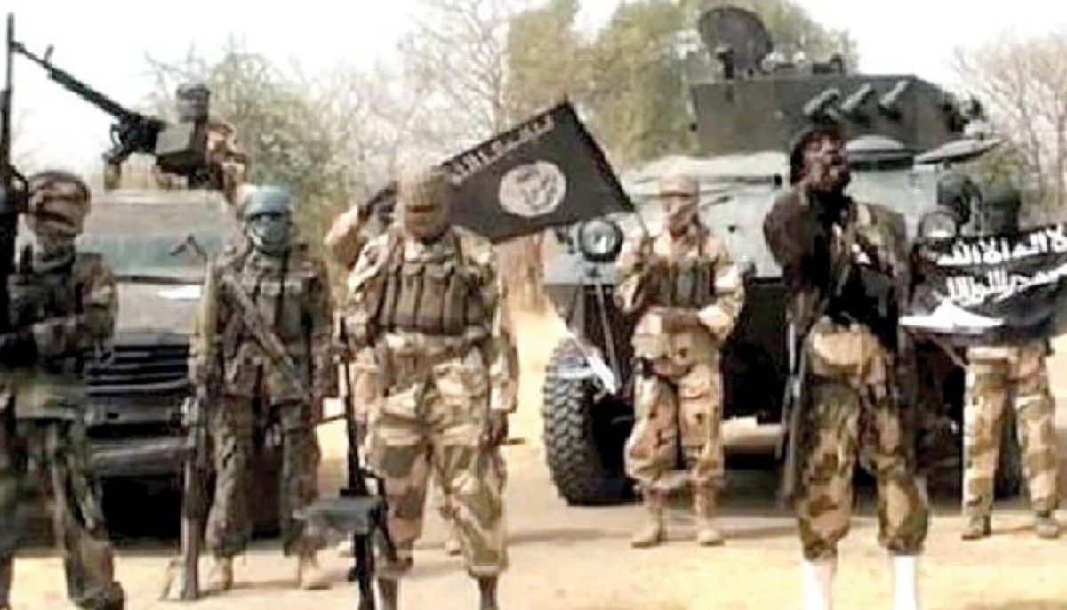 Group of armed militants in desert camouflage beside an armored vehicle, one raising a flag.