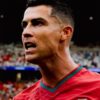Close-up of a male soccer player shouting on the field, wearing a red jersey with green trim.