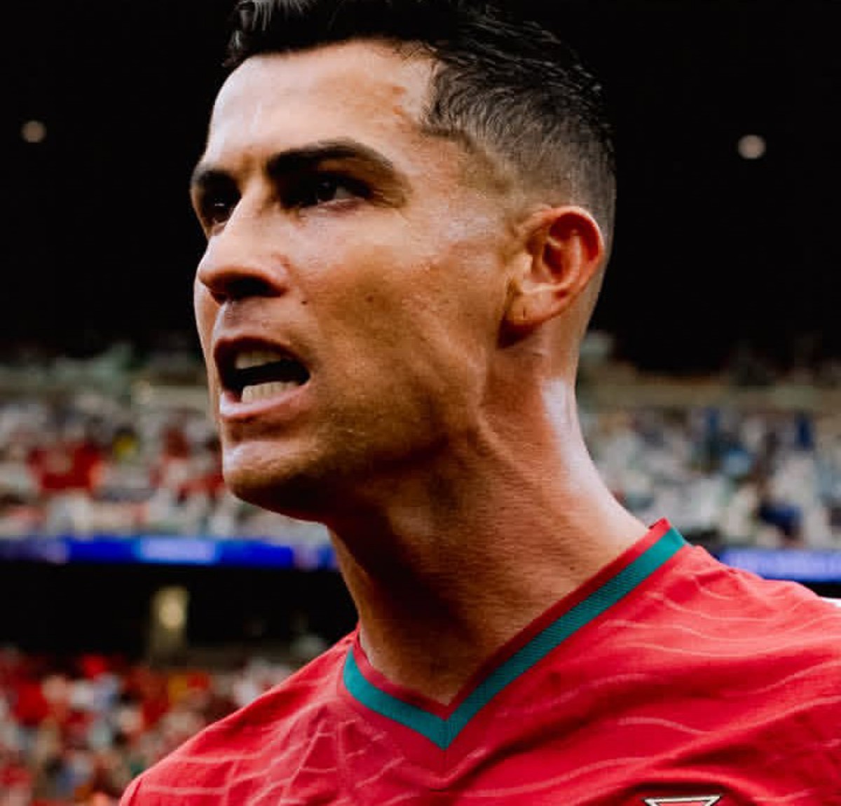 Close-up of a male soccer player shouting on the field, wearing a red jersey with green trim.