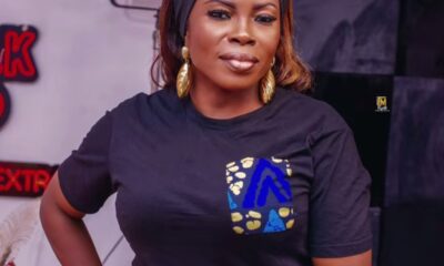 Woman with a headband and gold earrings, wearing a black T-shirt with a blue/yellow patterned pocket, posing with hand on hip.