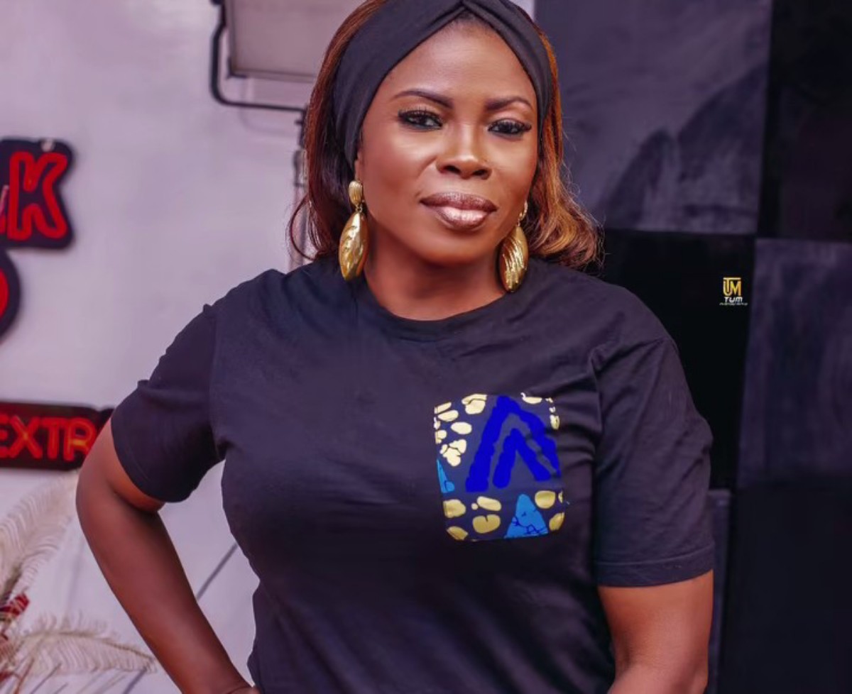 Woman with a headband and gold earrings, wearing a black T-shirt with a blue/yellow patterned pocket, posing with hand on hip.