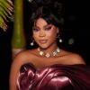 Glamorous portrait of a woman in an off‑the‑shoulder burgundy satin dress, wearing a pearl‑beam necklace and hoop earrings against a dark background.