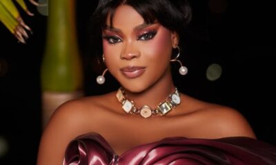 Glamorous portrait of a woman in an off‑the‑shoulder burgundy satin dress, wearing a pearl‑beam necklace and hoop earrings against a dark background.