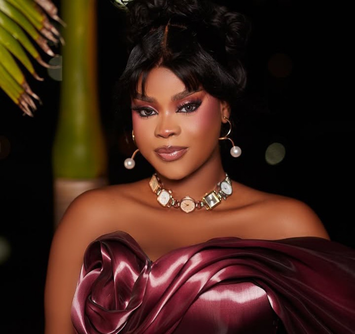 Glamorous portrait of a woman in an off‑the‑shoulder burgundy satin dress, wearing a pearl‑beam necklace and hoop earrings against a dark background.