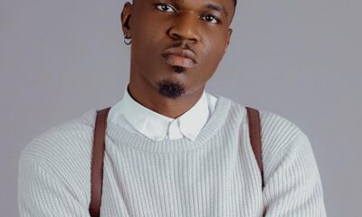 Portrait of a man with a trimmed goatee wearing a white knit sweater over a white collared shirt, looking at the camera against a gray backdrop with arms crossed.