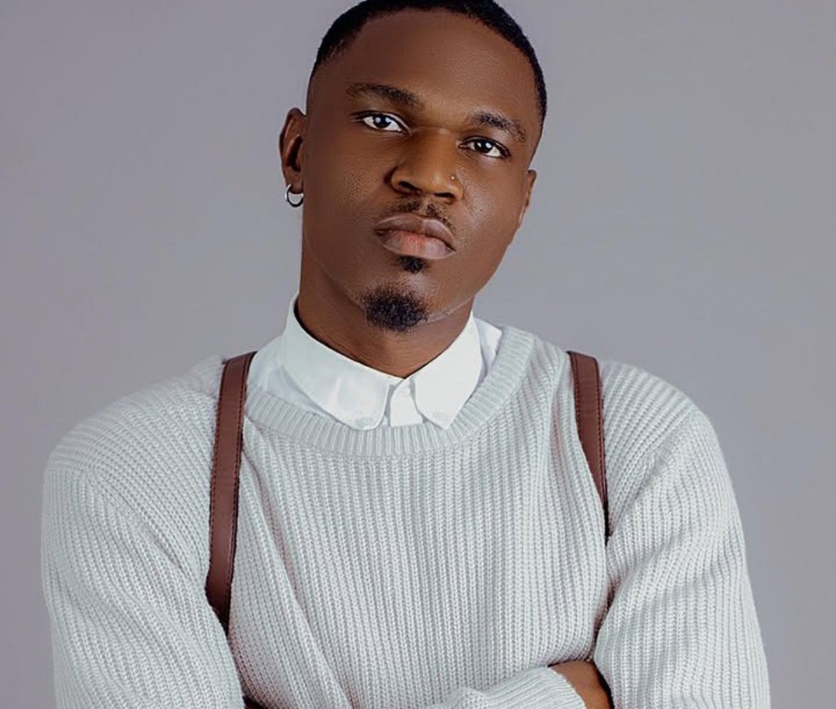 Portrait of a man with a trimmed goatee wearing a white knit sweater over a white collared shirt, looking at the camera against a gray backdrop with arms crossed.