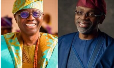 Two men smiling side by side: left in bright turquoise and gold traditional Nigerian attire with a patterned cap and bead necklace; right in blue medical scrubs with a red surgical cap.