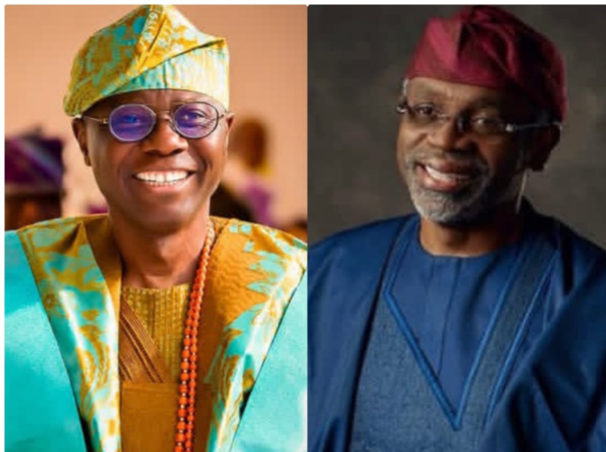 Two men smiling side by side: left in bright turquoise and gold traditional Nigerian attire with a patterned cap and bead necklace; right in blue medical scrubs with a red surgical cap.