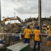 Construction workers in yellow safety vests and helmets stand among rubble as a yellow excavator clears debris at a collapsed site.